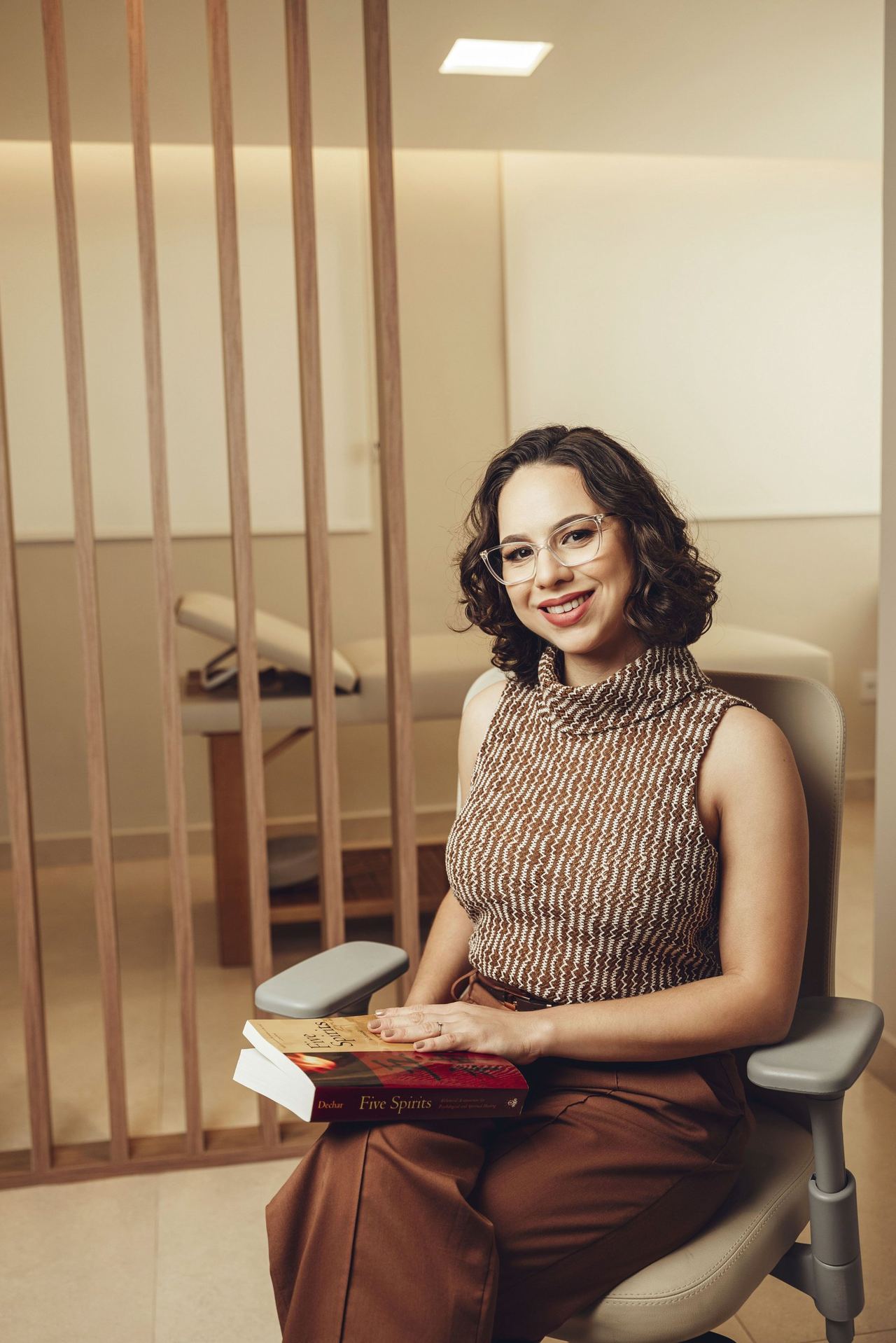 Dra. Thaís sentada em uma cadeira do consultório, com um livro no colo, olhando para a câmera.