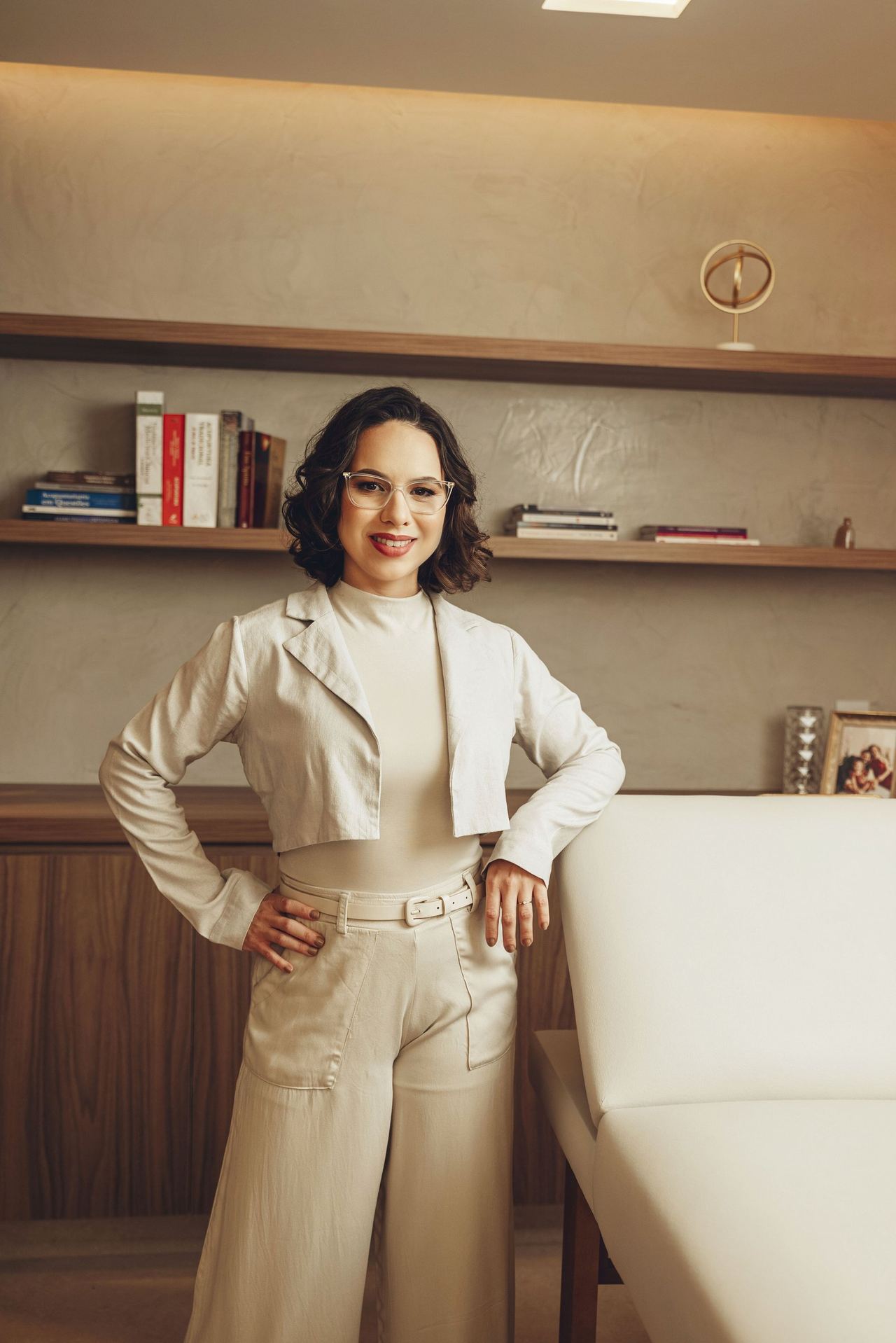 Dra. Thaís Santos em pé no consultório, diante de uma estante com livros.