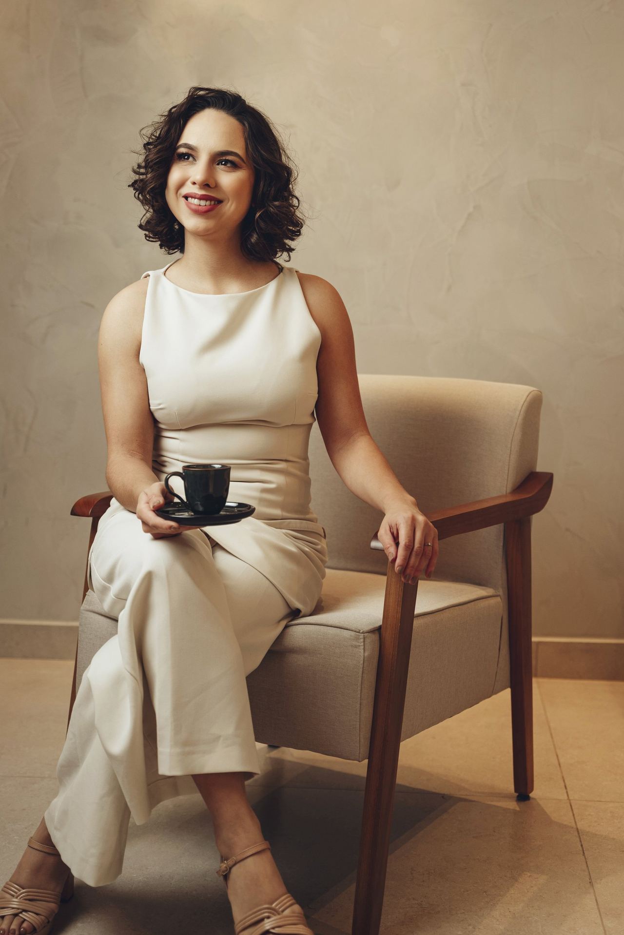 Dra. Thaís Santos sentada em uma poltrona do consultório, com uma xícara de café, sorrindo para a câmera.